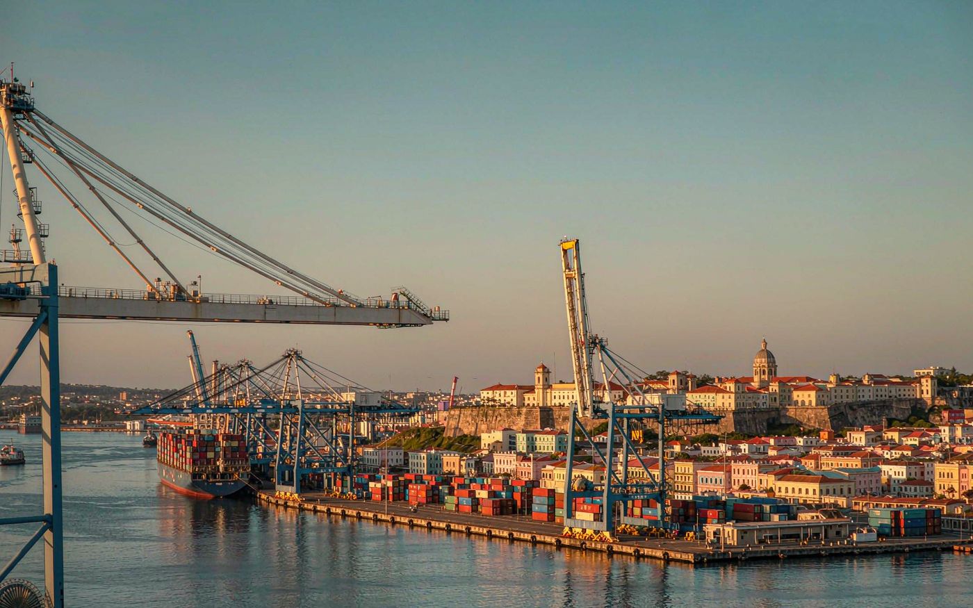 Wide photograph of a large Caribbean container port at golden hour with ship-to-shore gantry cranes loading a container ship, stacked containers in a paved yard, and the pastel colonial waterfront of Old San Juan with terracotta roofs and a stone fortress wall on the hill behind