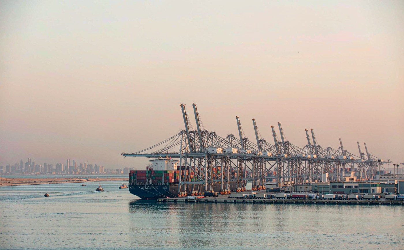 Container port on the Persian Gulf in soft morning haze, a modern deep water terminal with tall gantry cranes, a long straight quay, precise rows of stacked shipping containers, a large container ship berthed, flat desert coastline and a distant low city skyline