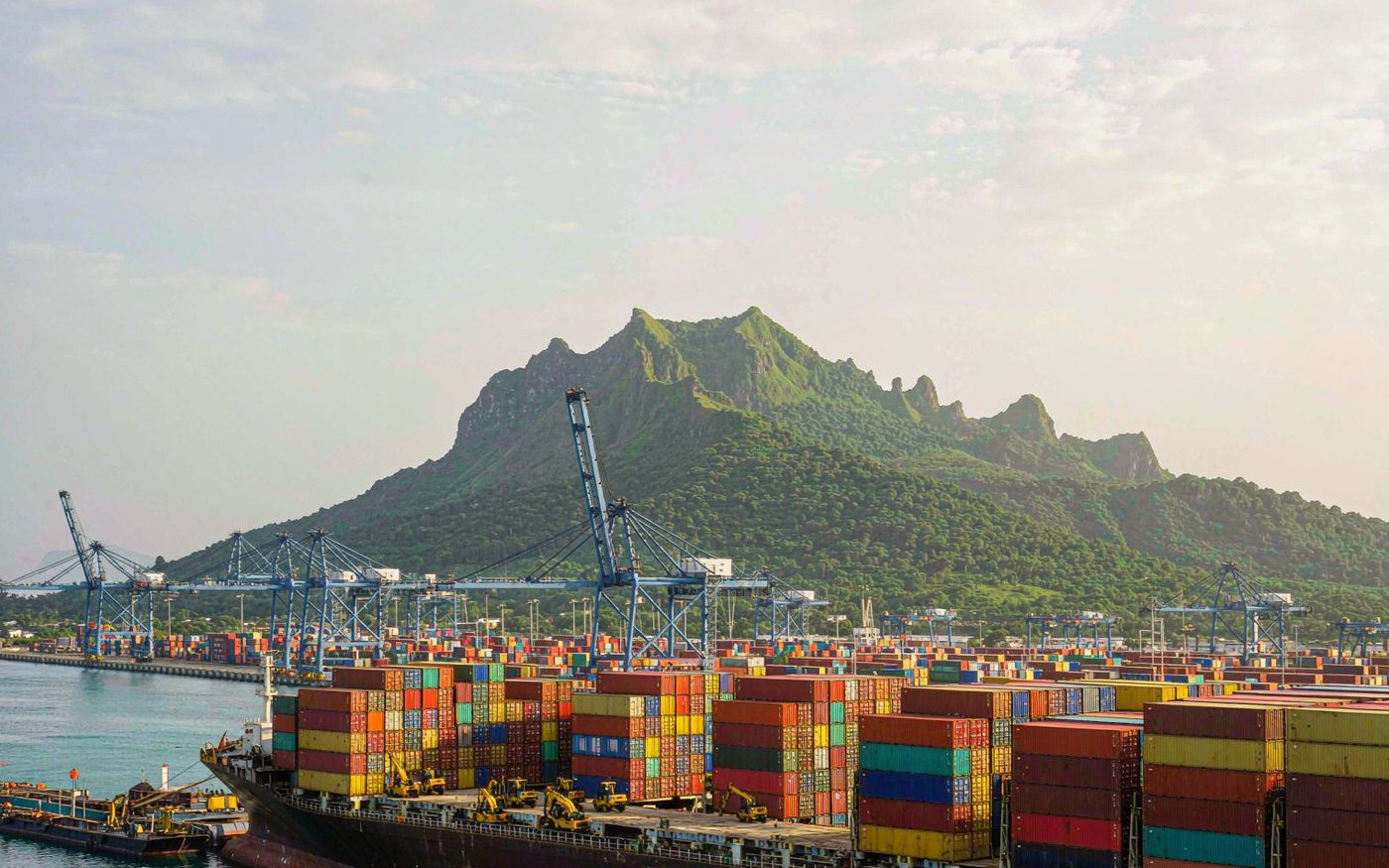 A container ship being worked by gantry cranes at a tropical Indian Ocean port with volcanic mountains behind