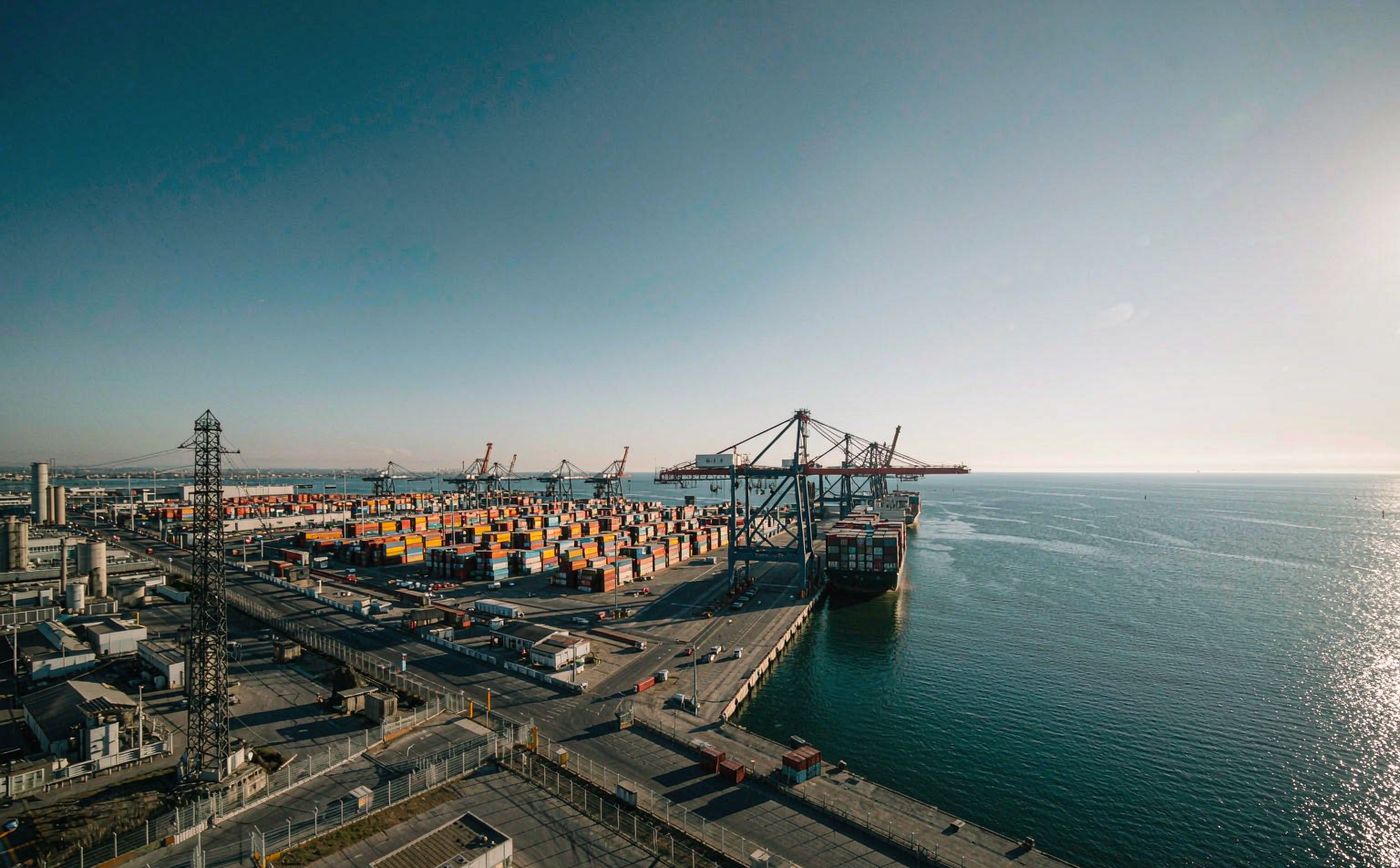 Black Sea container port in clear daylight, tall ship-to-shore gantry cranes, rows of stacked shipping containers in the yard, a container vessel at the quay and an open sea horizon