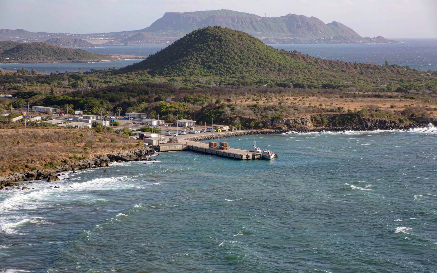 Tiny arid Dutch Caribbean island with a small concrete landing pier, one small cargo boat, a few pallets, dry scrubland and a rounded green volcano behind