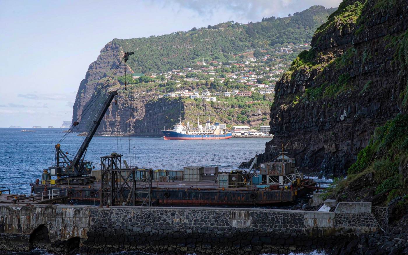 Narrow wharf at the foot of towering dark volcanic cliffs with a crawler crane lifting cargo and a flat-top lighter barge alongside, a supply ship anchored offshore in an open roadstead, a small town in a steep green valley, deep blue South Atlantic water under clear island light