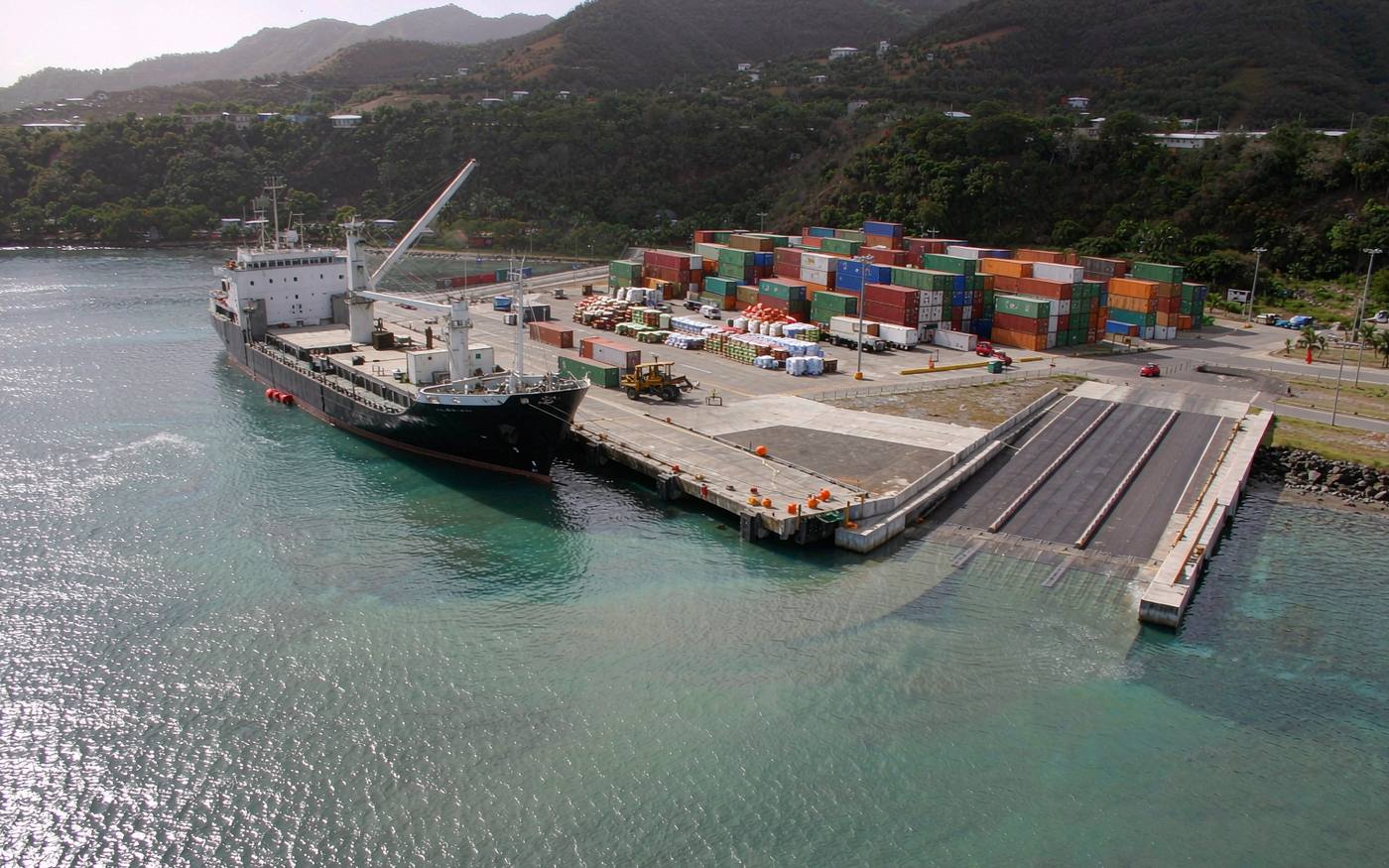 Small Caribbean harbour with a concrete cargo pier, a feeder vessel alongside, crated cargo on the quay and green volcanic hills behind