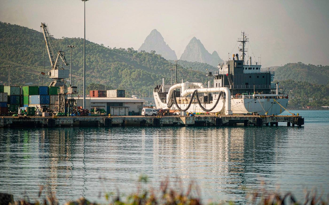 Caribbean container quay with a small gantry crane, stacked containers, a berthed cargo vessel and green mountains hazy in the distance