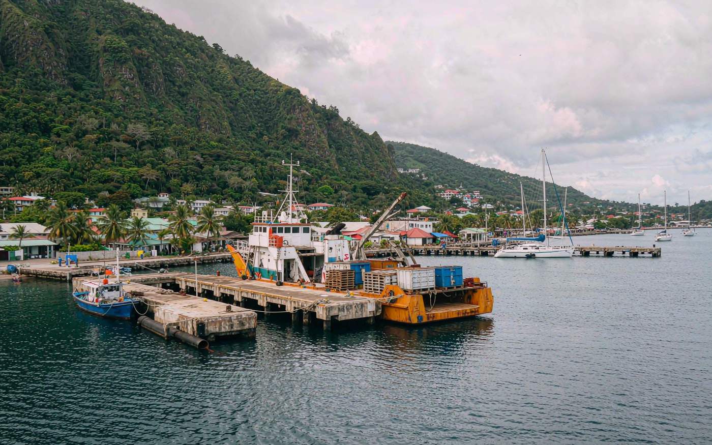 Small Caribbean waterfront harbour with a short concrete pier, an inter-island cargo vessel unloading pallets, steep green slopes behind and yachts anchored offshore