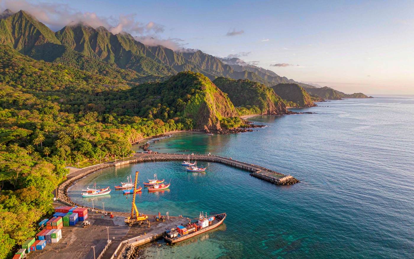 Aerial documentary view of a small container quay on the volcanic island of Sao Tome in the Gulf of Guinea, a few stacked shipping containers and a mobile harbour crane, dense rainforest on steep green peaks behind, turquoise Atlantic water and a breakwater, warm equatorial afternoon light