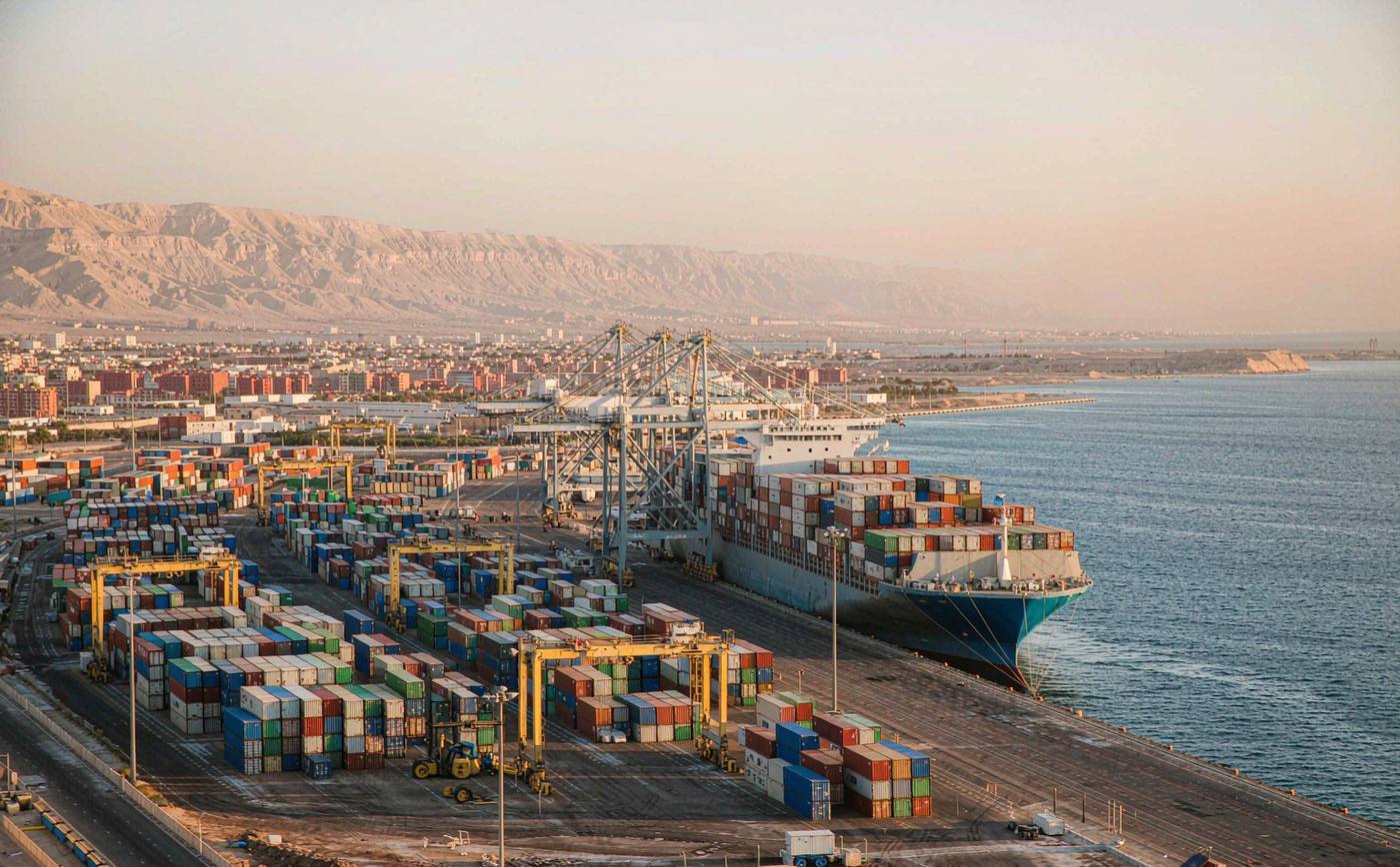 Container port on the Red Sea in late afternoon light, gantry cranes and straddle carriers working a long quay, neat blocks of stacked shipping containers, a large container vessel alongside, pale arid mountains and a low port city skyline behind