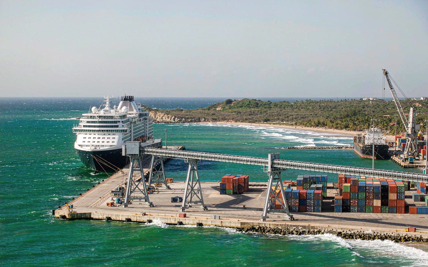 Wide photograph of a Caribbean island port from the water with a cruise and cargo quay, stacked containers in a working yard, low-rise brightly coloured waterfront buildings and a hillside town behind, with turquoise water in the foreground