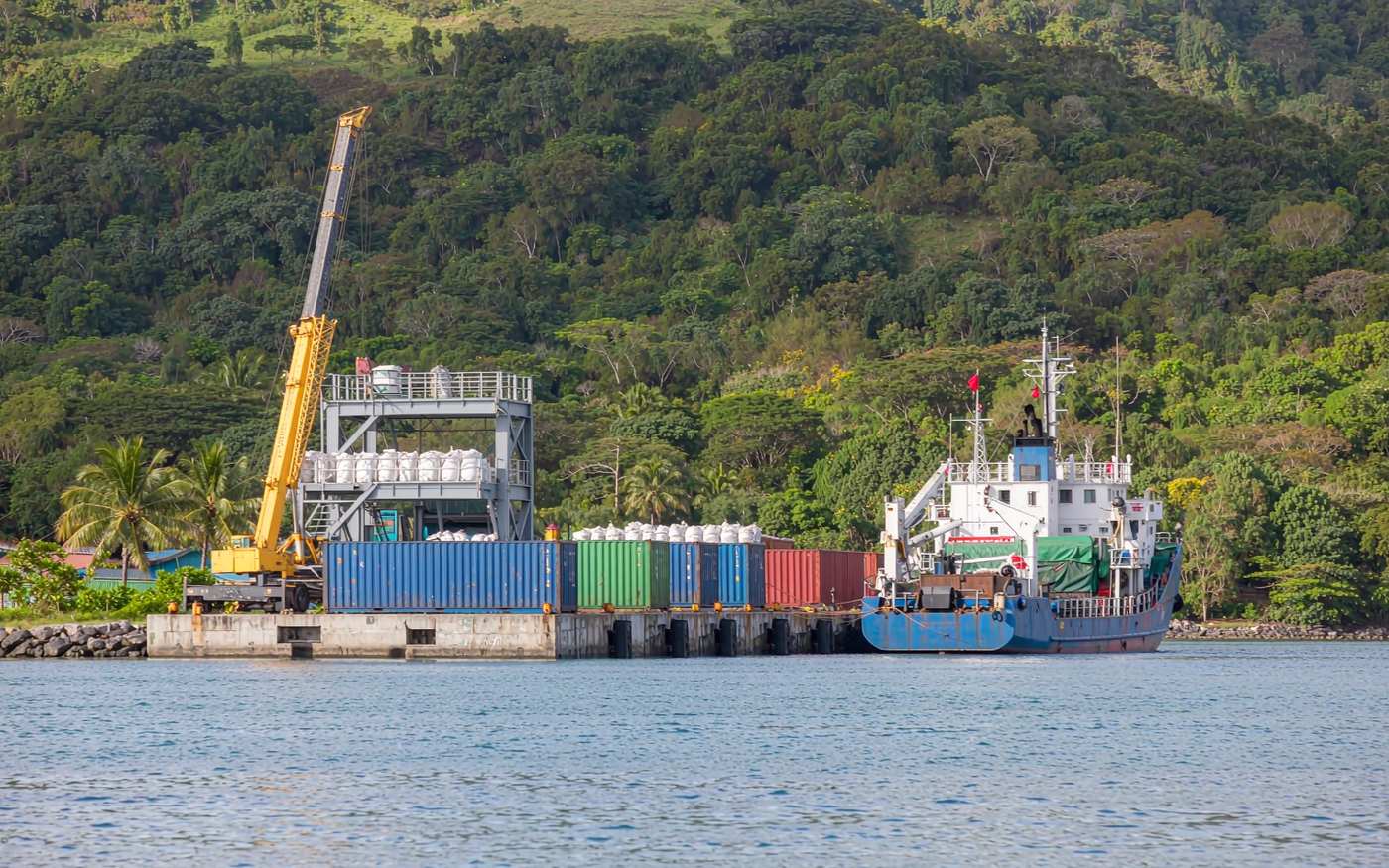 Small Melanesian container quay with a mobile crane, stacked containers and bagged cargo, a coastal cargo vessel alongside and densely forested green hills behind