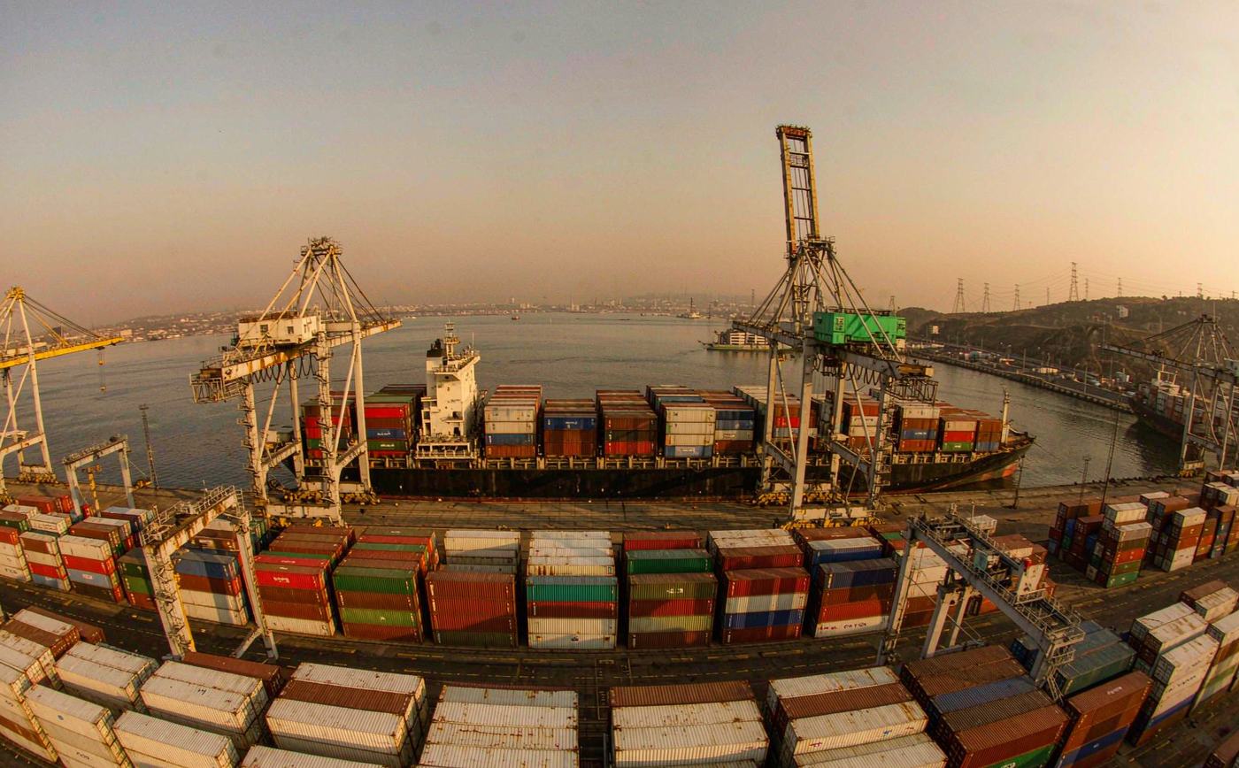 A large South African container terminal in warm late afternoon light, rows of stacked shipping containers, tall gantry cranes, a container vessel berthed at the quay and industrial port infrastructure
