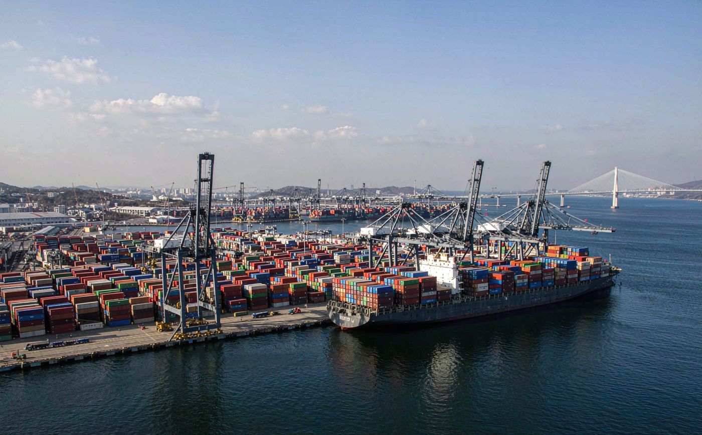 Container terminal in Busan, South Korea in clear daylight, a busy deep water quay with tall gantry cranes and dense rows of stacked shipping containers, a large container ship berthed alongside, a bridge and cranes in the distance, calm blue harbour water