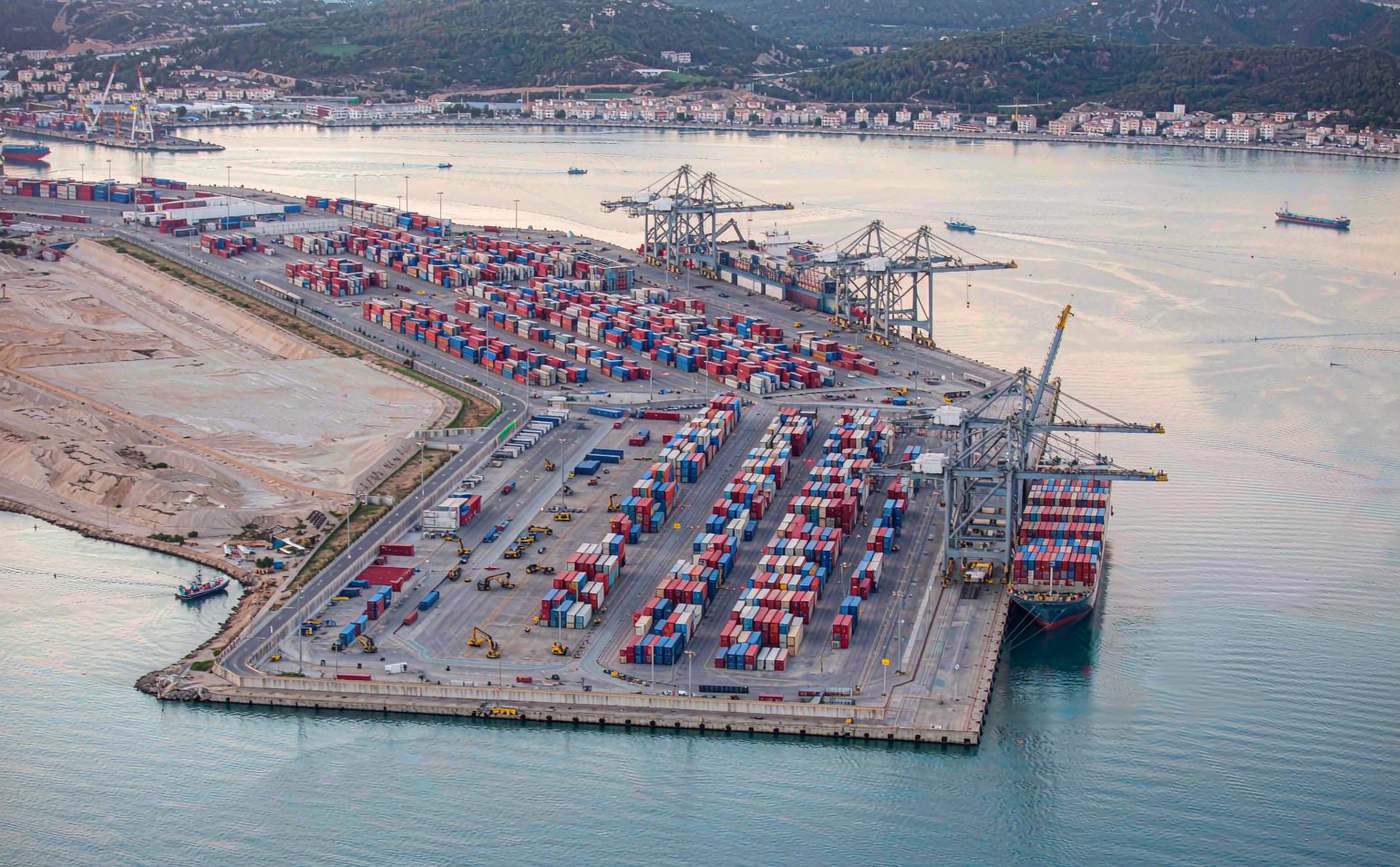 Mediterranean container port with ship-to-shore gantry cranes, rows of stacked containers and a large vessel berthed at the quay