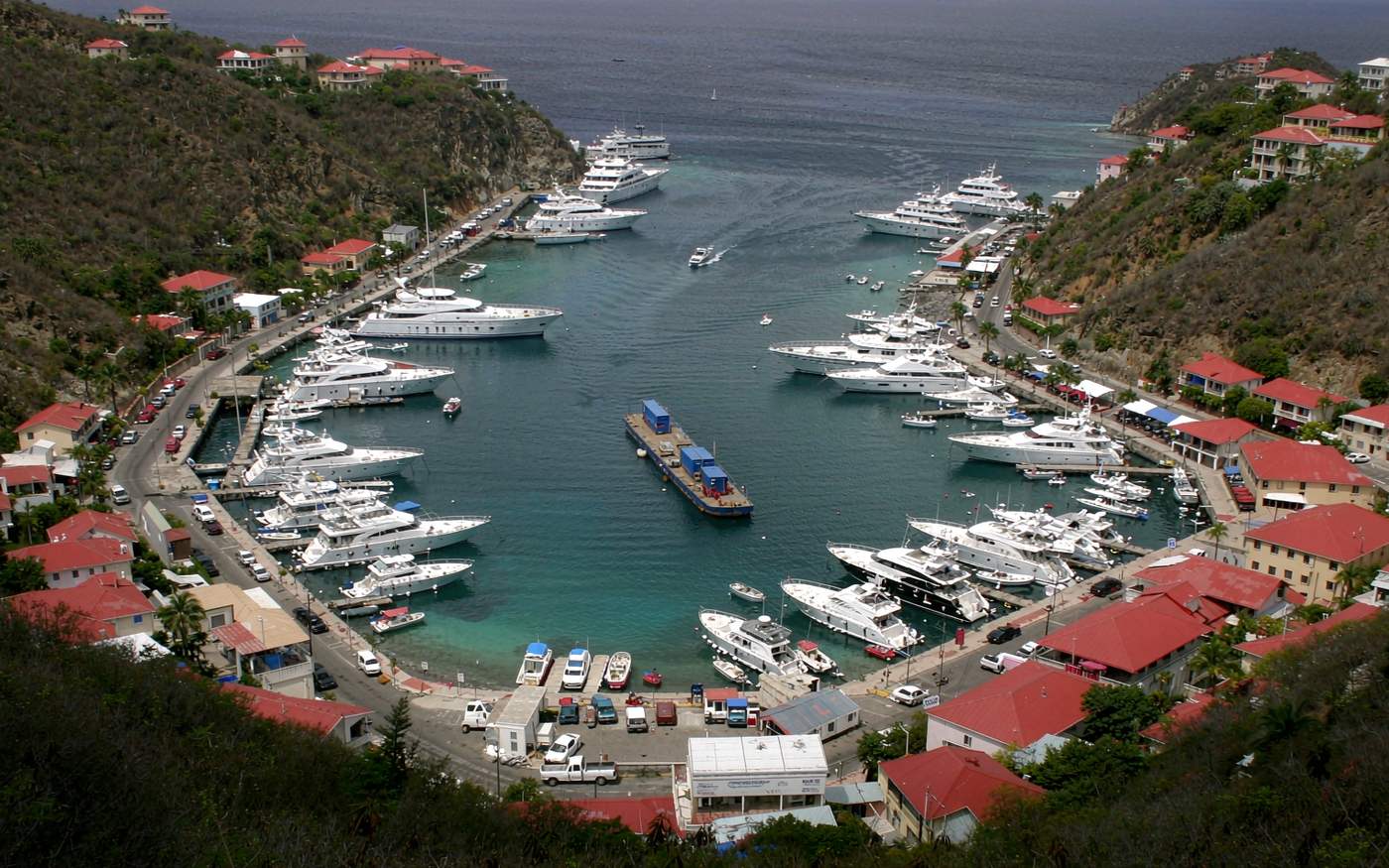 Documentary photograph of the small horseshoe harbour at Gustavia in Saint Barthelemy, red-roofed colonial buildings and luxury yachts lining the quay, a small cargo barge and a tender boat at the waterfront, steep arid green hills framing the bay, turquoise Caribbean water under bright tropical daylight