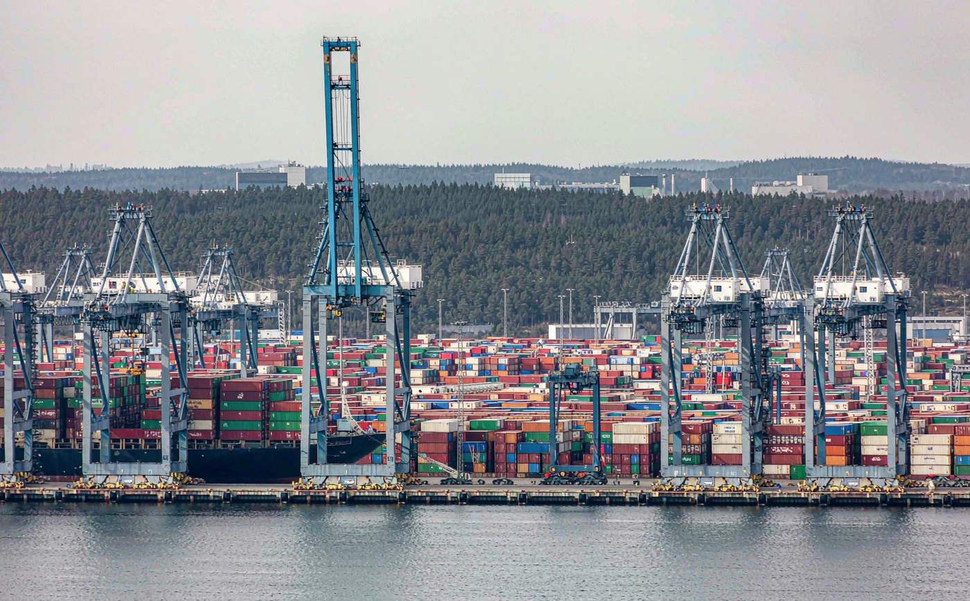 Large container terminal with tall gantry cranes at the quay, a container vessel being loaded, stacked containers in the yard and low Scandinavian pine hills behind