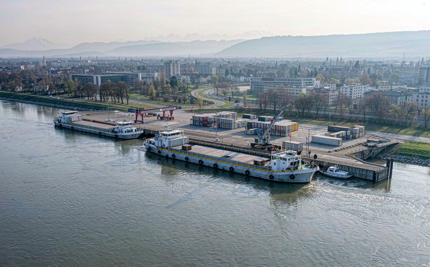 River port terminal on the Rhine with container barges at the quay, a harbour crane loading containers, stacked containers in the yard and low Alpine foothills in the distance