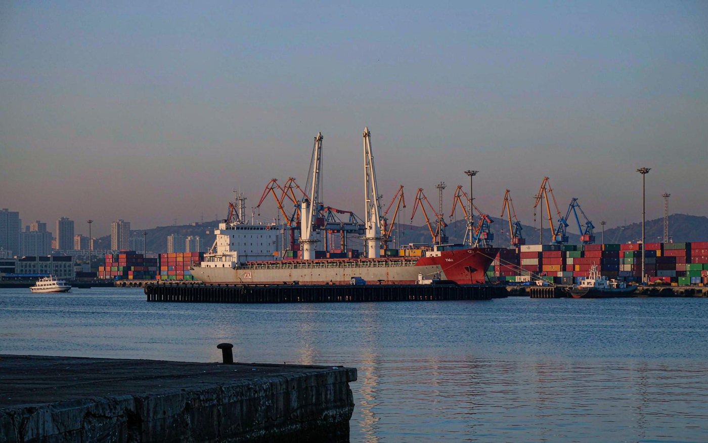 Photograph of an eastern Mediterranean container port in the early morning with a mid-size cargo ship at berth, container stacks and portal cranes, calm blue water and low green hills behind the city