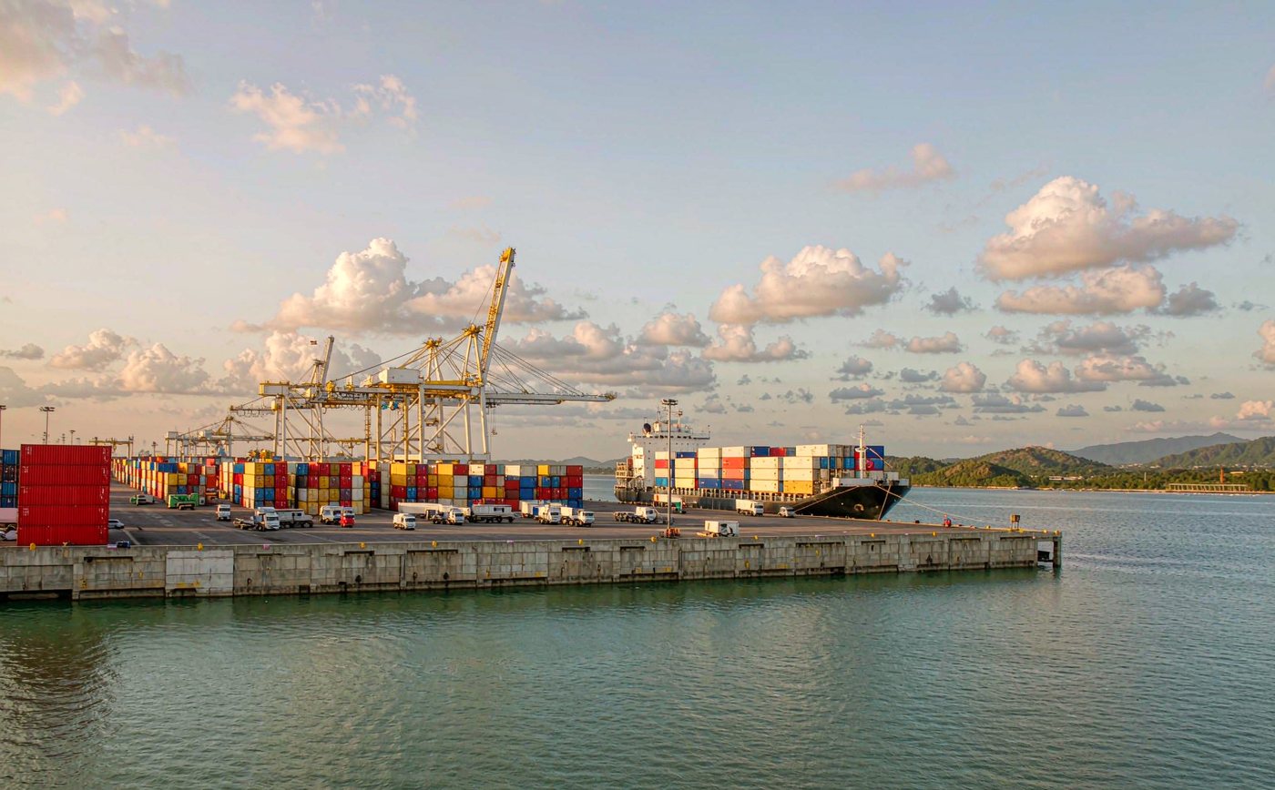 A deep water container terminal on the Gulf of Thailand in late afternoon light, rows of stacked shipping containers, two tall gantry cranes, a container vessel at berth, calm green blue water, low green hills and tropical clouds on the horizon