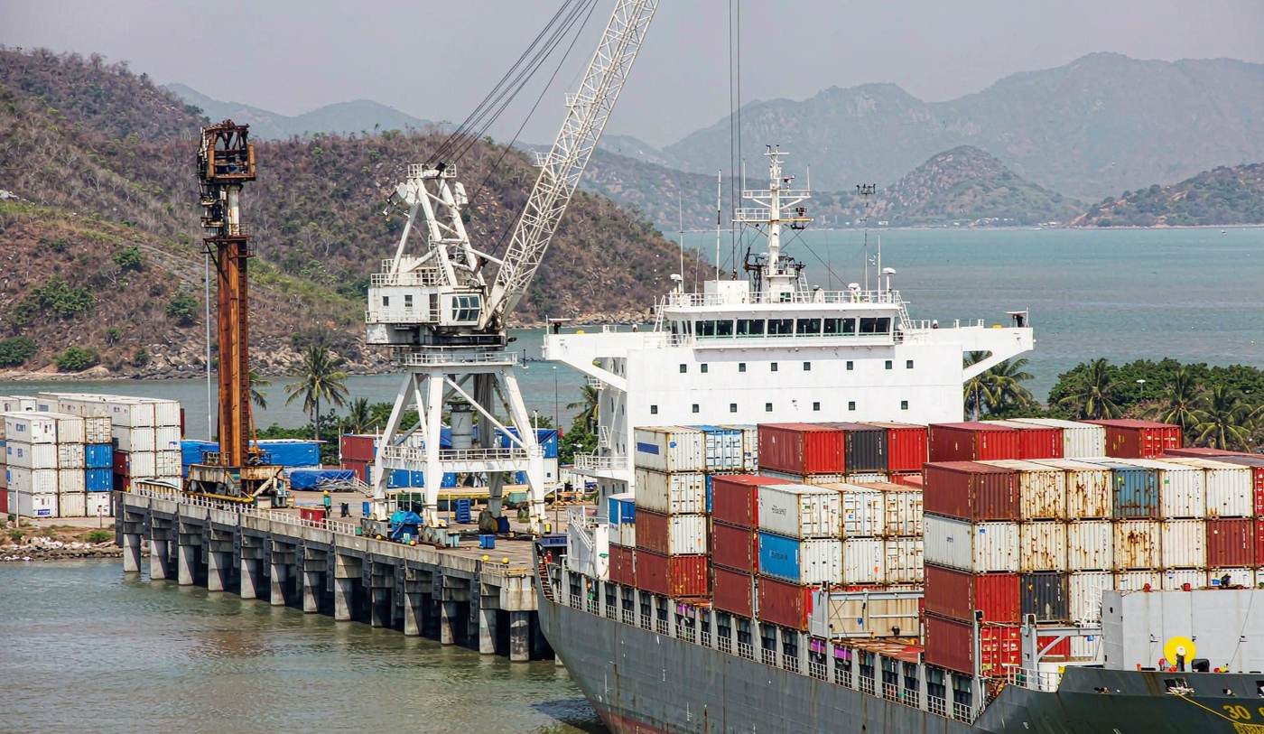 A small deep-water container port on a tropical island with a container vessel alongside a modern quay, a shore crane and stacks of shipping containers, dry green hills and a wide bay behind under bright hazy tropical sunlight
