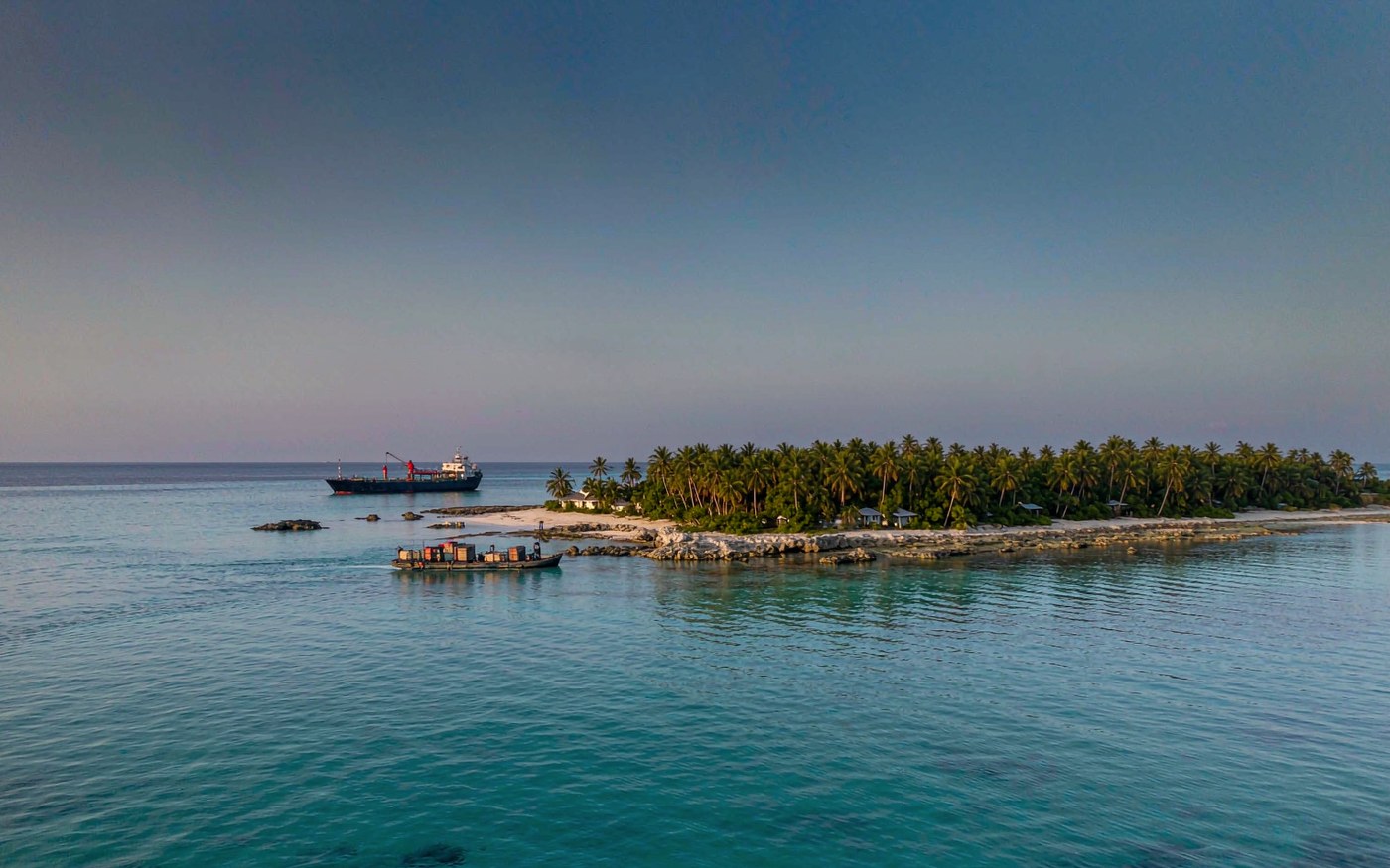 Low coral atoll in the South Pacific with a small cargo vessel anchored offshore and a work boat ferrying crates to the beach