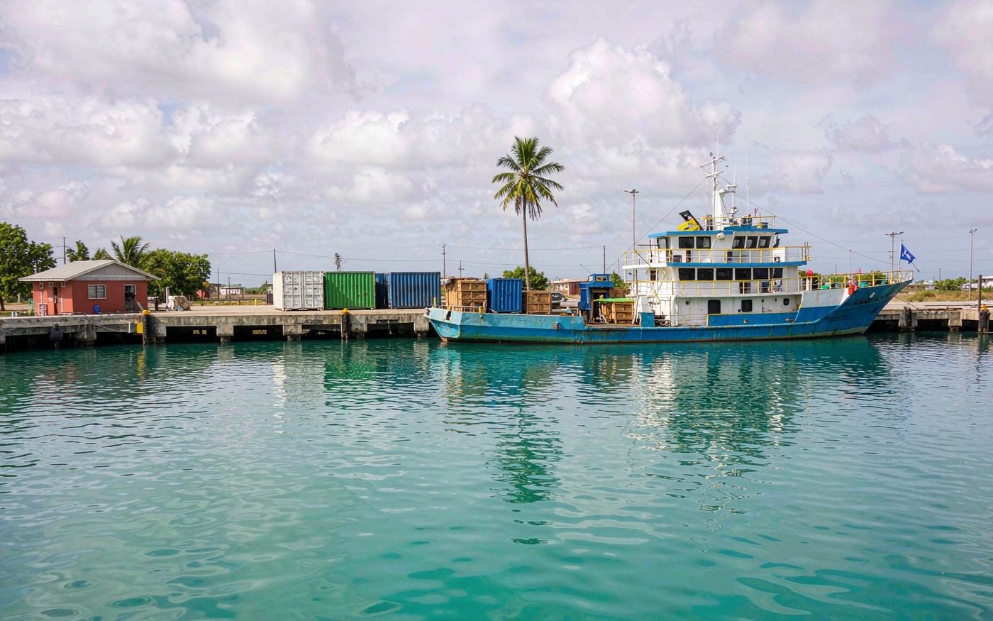 Low coral island port with a concrete berth, an inter-island cargo vessel unloading crates, stacked containers on the quay, palm trees and turquoise lagoon water