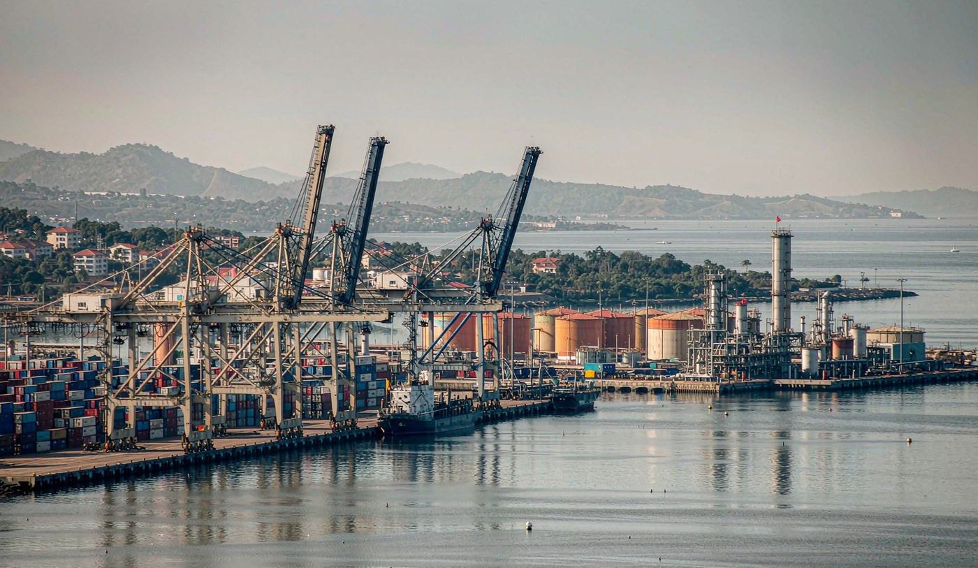 A large commercial container port in the southern Caribbean with gantry cranes over stacked shipping containers, a container vessel at berth and industrial petrochemical storage tanks in the middle distance