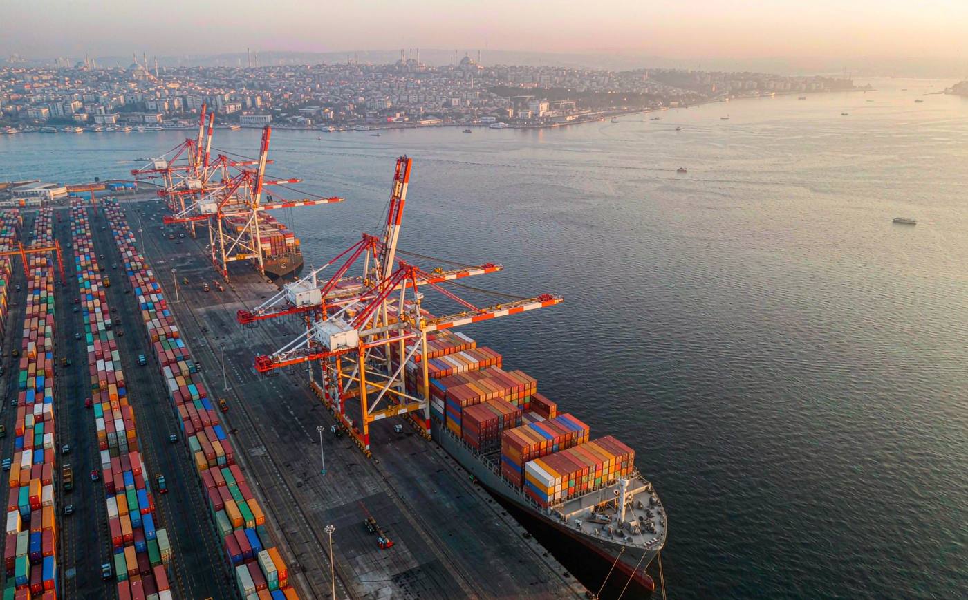 Aerial view of a container terminal on the Sea of Marmara near Istanbul in late afternoon light, gantry cranes over a long quay, dense rows of shipping containers, a container vessel berthed and a hazy city skyline with domes and minarets far behind