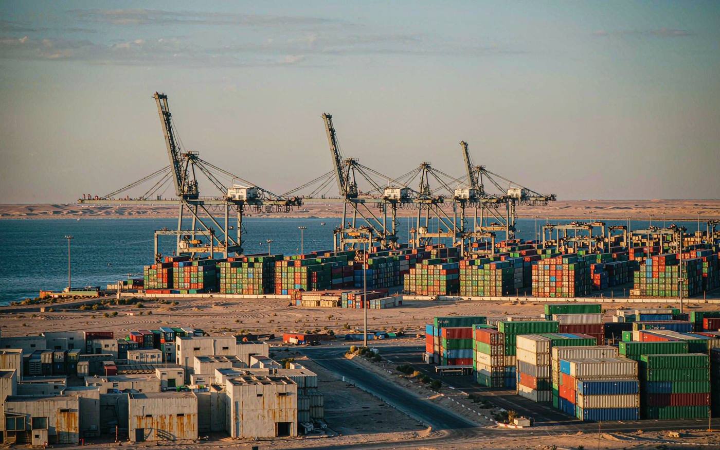 Photograph of a container terminal on the Caspian Sea at late afternoon with stacked shipping containers in green blue and red, gantry cranes, calm water and a flat desert coastline