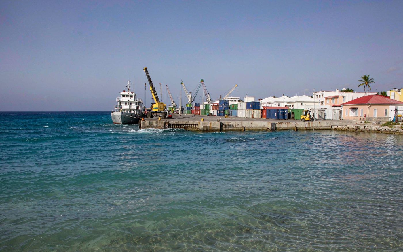 Photograph of a small Caribbean island working port with vivid turquoise shallow water, a modest container yard with stacked boxes and a mobile harbour crane on a concrete quay, low-rise white and pastel buildings behind and a small coaster alongside