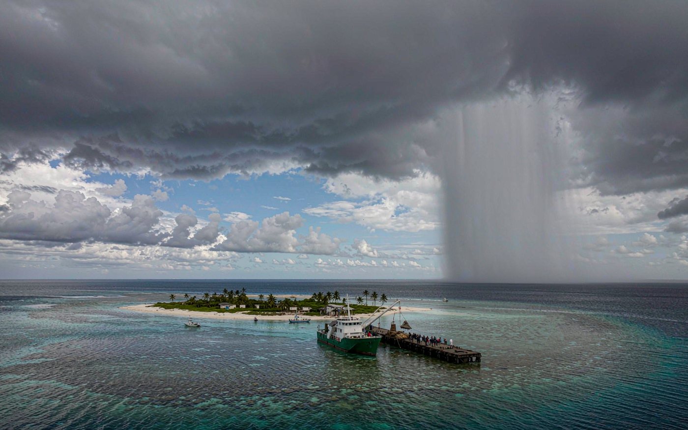 Very small coral atoll whose wharf sits barely above sea level, a feeder ship discharging cargo ashore, small boats, coastal palms and storm clouds above