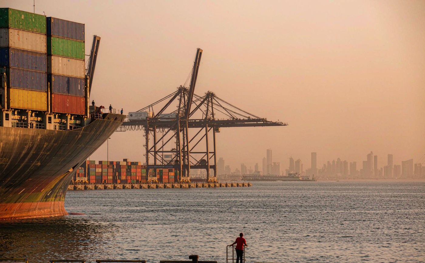 Container terminal on the Arabian Gulf at first light, gantry cranes loading a large container ship, long rows of stacked shipping containers, calm water and a hazy modern skyline far behind