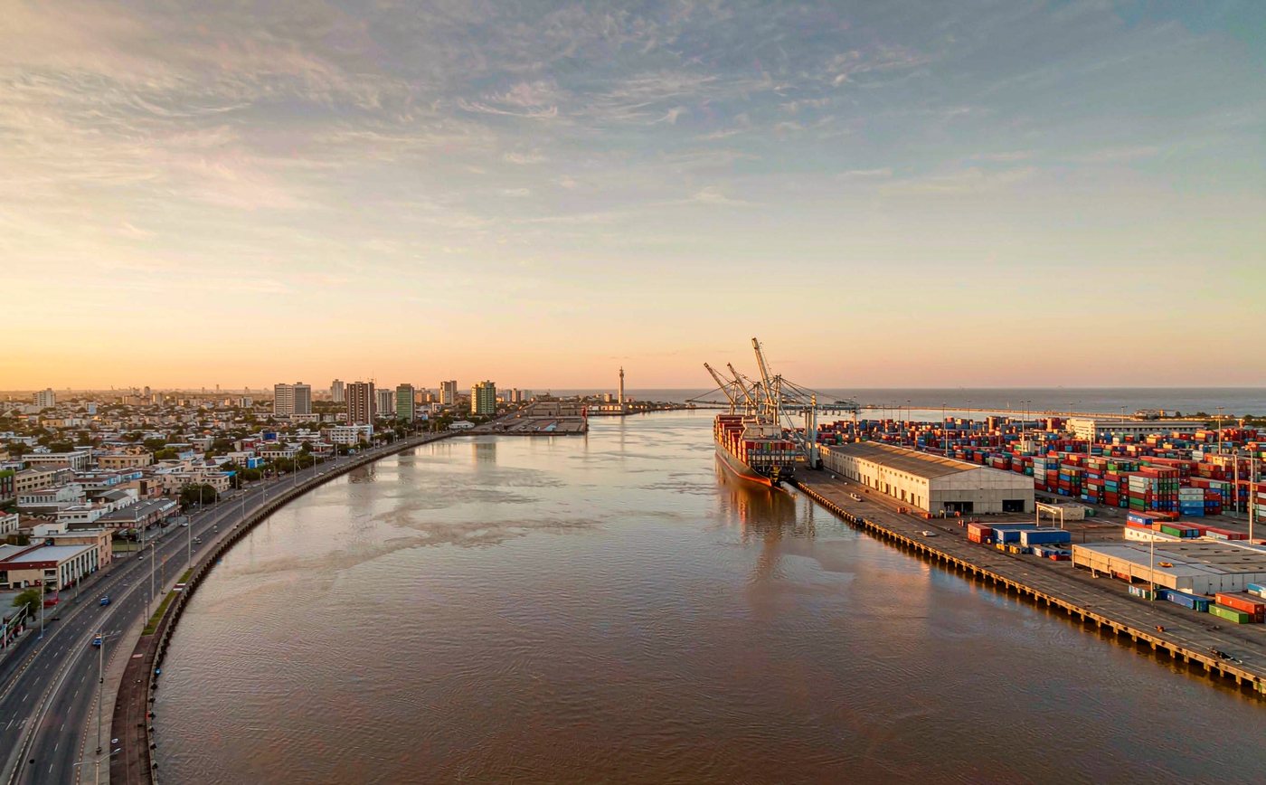 The container terminal at the Port of Montevideo on the broad brown estuary of the River Plate in late afternoon light, with a container ship at the quay, gantry cranes, stacked containers, a low city skyline and calm water to the horizon