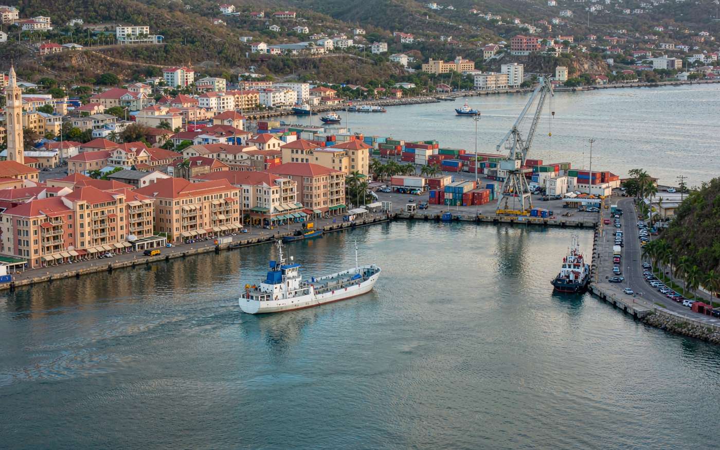 Elevated photograph of a Caribbean harbour town with red-roofed colonial waterfront buildings along a deep natural bay, a working cargo quay with stacked shipping containers and a small gantry crane, a coaster and a tug in the water and green hills behind