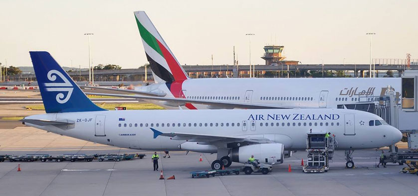 Air New Zealand wide-body aircraft connected to a jet bridge at a Chinese airport, with an Emirates tail visible behind the terminal
