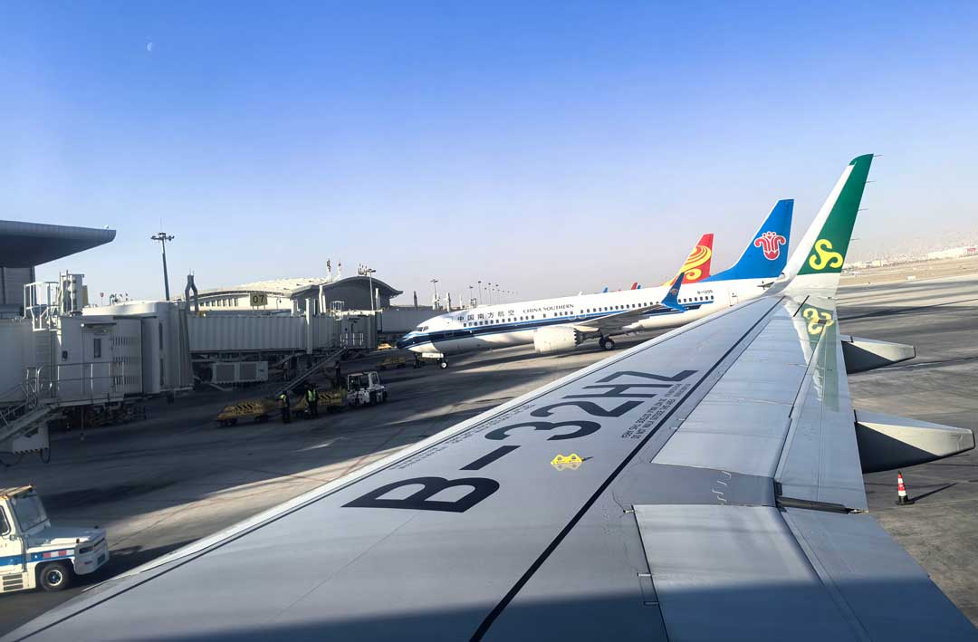 Aircraft wing seen from a window seat over the apron of a Chinese airport, with a China Southern aircraft and jet bridges in the background
