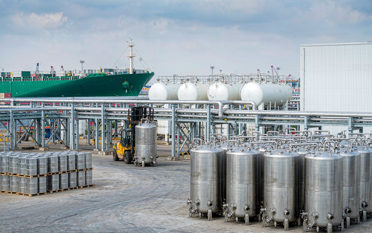Liquid chemical terminal in China with rows of steel drums and stainless steel tanks under pipework, and a tanker vessel berthed behind