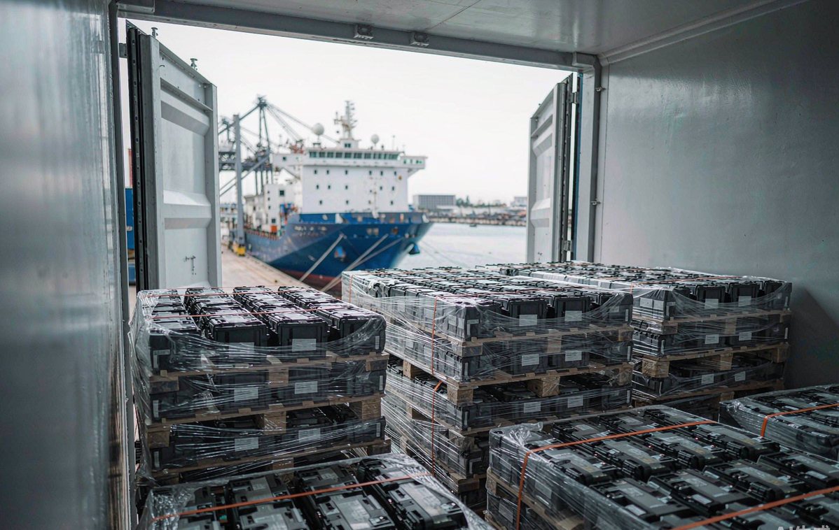 Palletised lithium battery packs shrink-wrapped inside a shipping container at a Chinese logistics hub, with a container vessel visible beyond the container doors