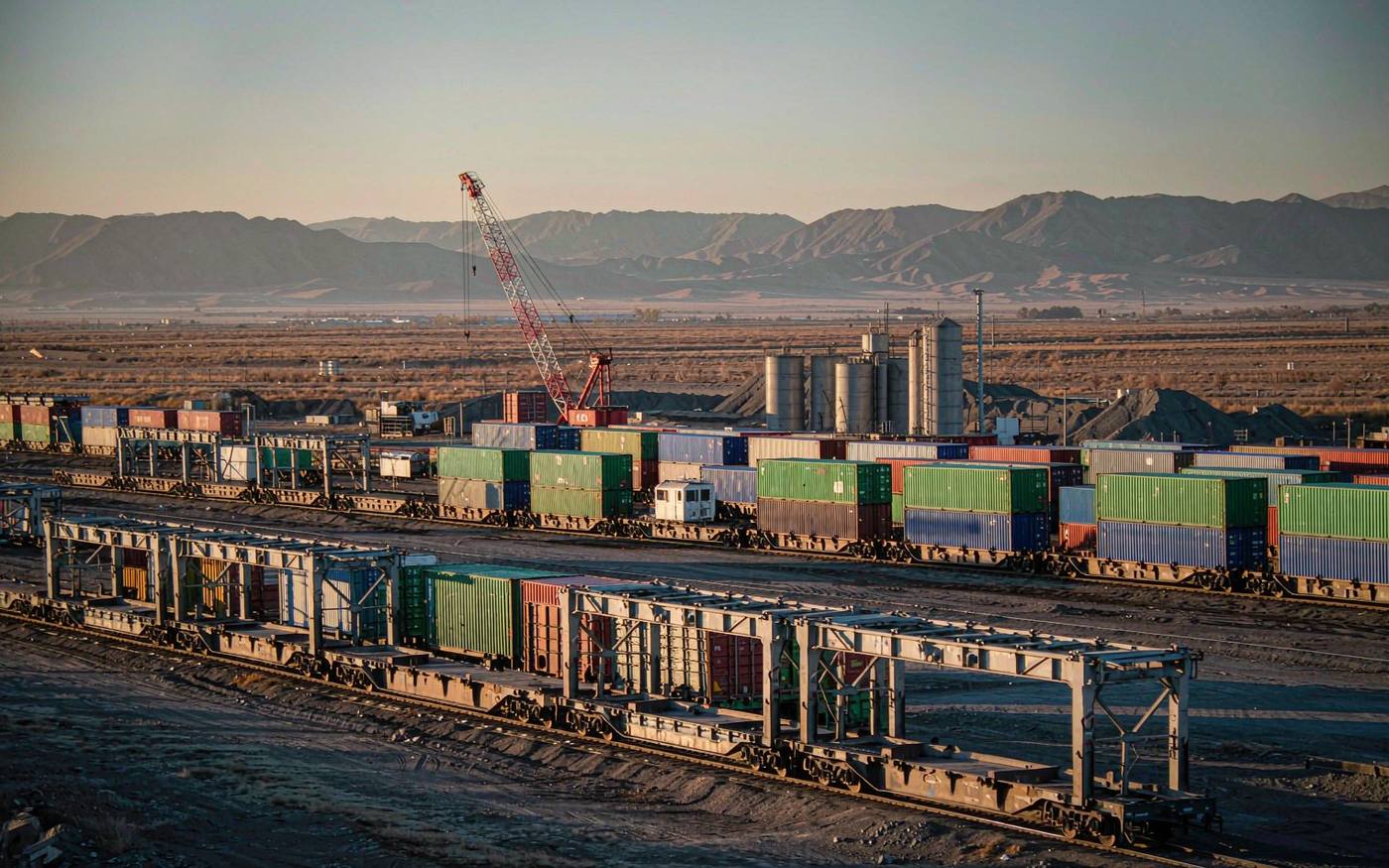 Photograph of a rail container terminal in Central Asia at late afternoon with intermodal freight trains, stacked containers in green blue and red, gantry cranes and distant mountains