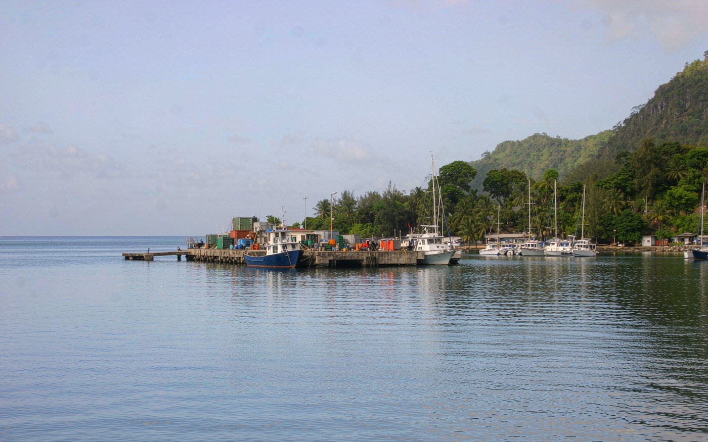 Small South Pacific harbour with a working wharf, stacked containers, a cargo vessel alongside, sailing yachts moored in the bay and steep green volcanic hills behind