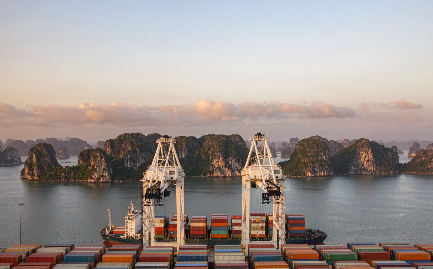 A modern Vietnamese deep water container terminal at golden hour, long rows of stacked shipping containers, two tall gantry cranes, a container vessel alongside the quay, calm harbour water and limestone karst islands on the horizon
