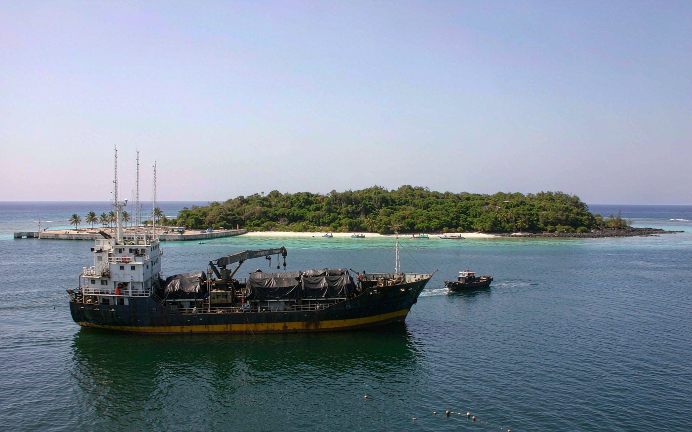 Cargo vessel anchored off a low green South Pacific island with a small work boat moving between the ship and the shore