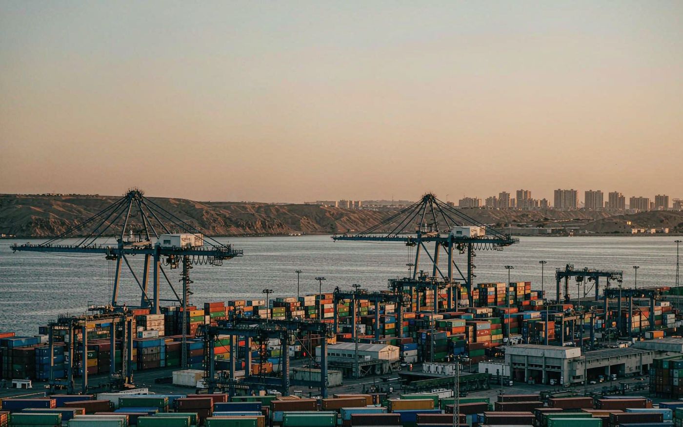 Photograph of a container terminal on the Red Sea coast at golden hour with stacked shipping containers in green blue and red, gantry cranes, calm harbour water and arid hills behind the port