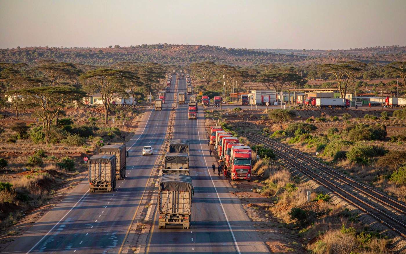 A truck convoy carrying shipping containers on a highway across dry African savanna with a rail line alongside