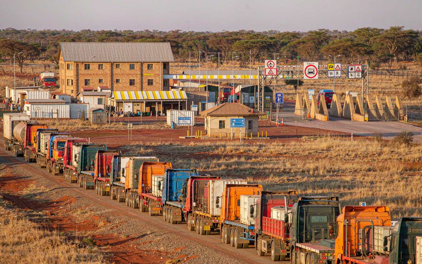 A queue of container trucks at a busy southern African border crossing with customs buildings and dry grassland